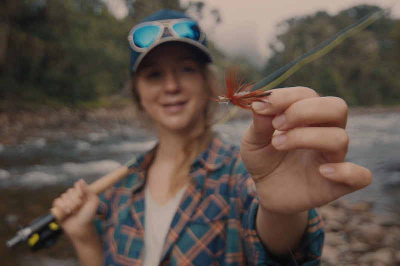 A woman in a blue plaid shirt and cap holds up a fishing fly lure with a green tail and orange-brown body, with a blurred river and forest background.