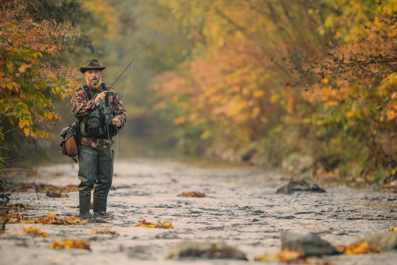 A man stands waist-deep in a flowing stream, fly fishing with a rod and reel. He is wearing a wide-brimmed hat, camouflage top, dark pants, and waders, surrounded by autumn trees with yellow and orange foliage.