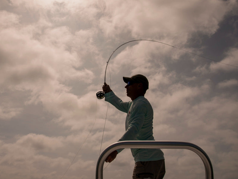 A silhouetted person fishing from a boat, holding a bent fishing rod, against a dramatic cloudy sky.